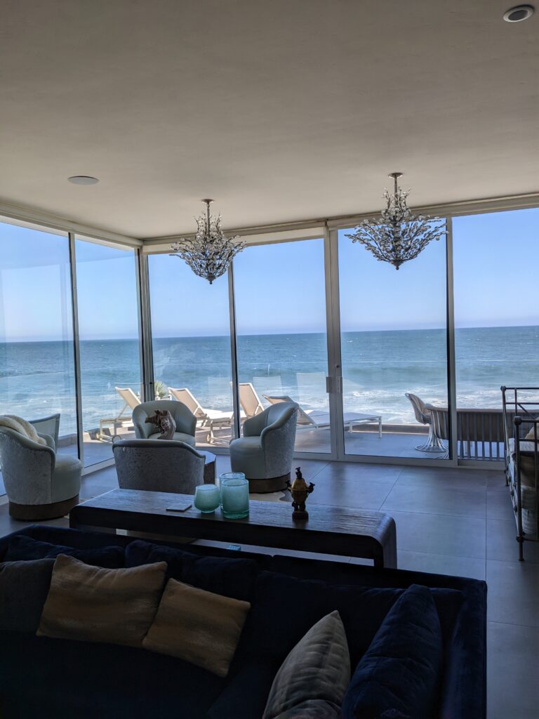 Malibu beach house with room showing two chandeliers, a couch, four light blue chairs, and a glassed in balcony showing the blue waters of Malibu