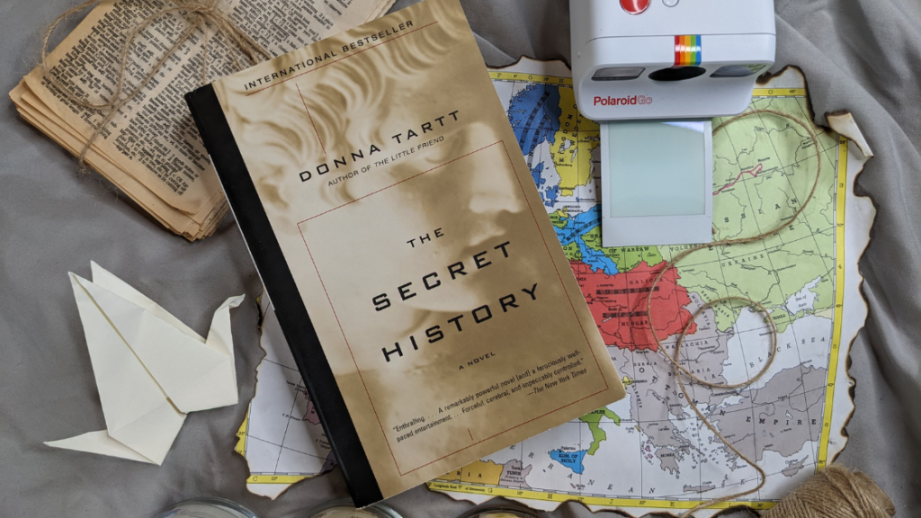 The Secret History (one of the books on the list) with an origami crane, a brunt map, an instant camera about to develop a pictures, twine, and yellowed dictionary pages